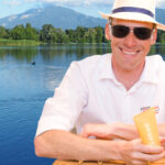 Mann mit Sonnenhut, Sonnenbrille, Hemd vor einem See, Sonnencreme in der Hand
