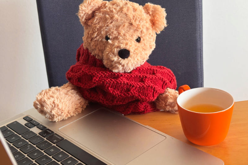 Teddy sitzt vor dem Computer, hat einen Schal um den Hals gewickelt und eine Tasse Tee vor sich am Tisch stehen