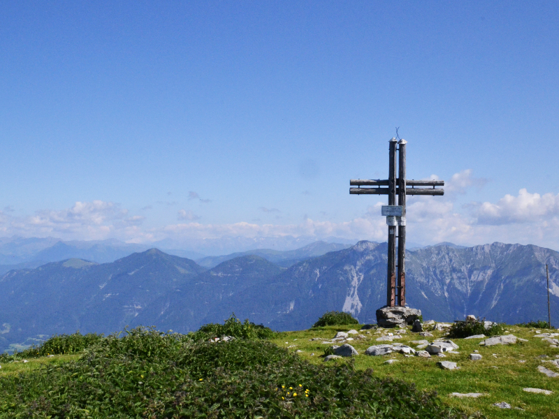 Berggipfel mit Gipfelkreuz (Poludnig), dahinter begrenzt eine Bergkette den Horizont.