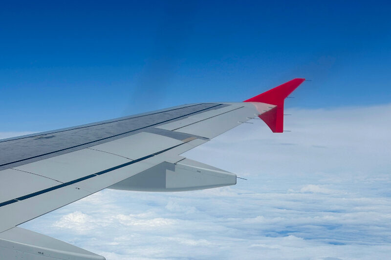 Flugzeugflügel, dahinter oben blauer Himmel, im unteren Bildbereich weiße Wolken.