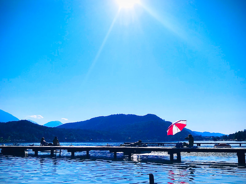 Blick auf einen Badesteg am Klopeiner See mit rot-weißem Sonnenschirm bei strahlendem Sonnenschein