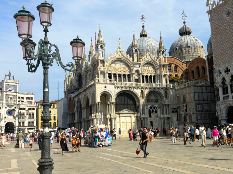 Markusplatz in Venedig mit der Kirche im Hintergrund, Laterne im vorderen Bereich des Bildes und man sieht einige Menschen auf dem Platz flanieren.