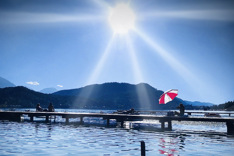 Steg am Klopeinersee mit rot-weißem Sonnenschirm und intensiver Sonne